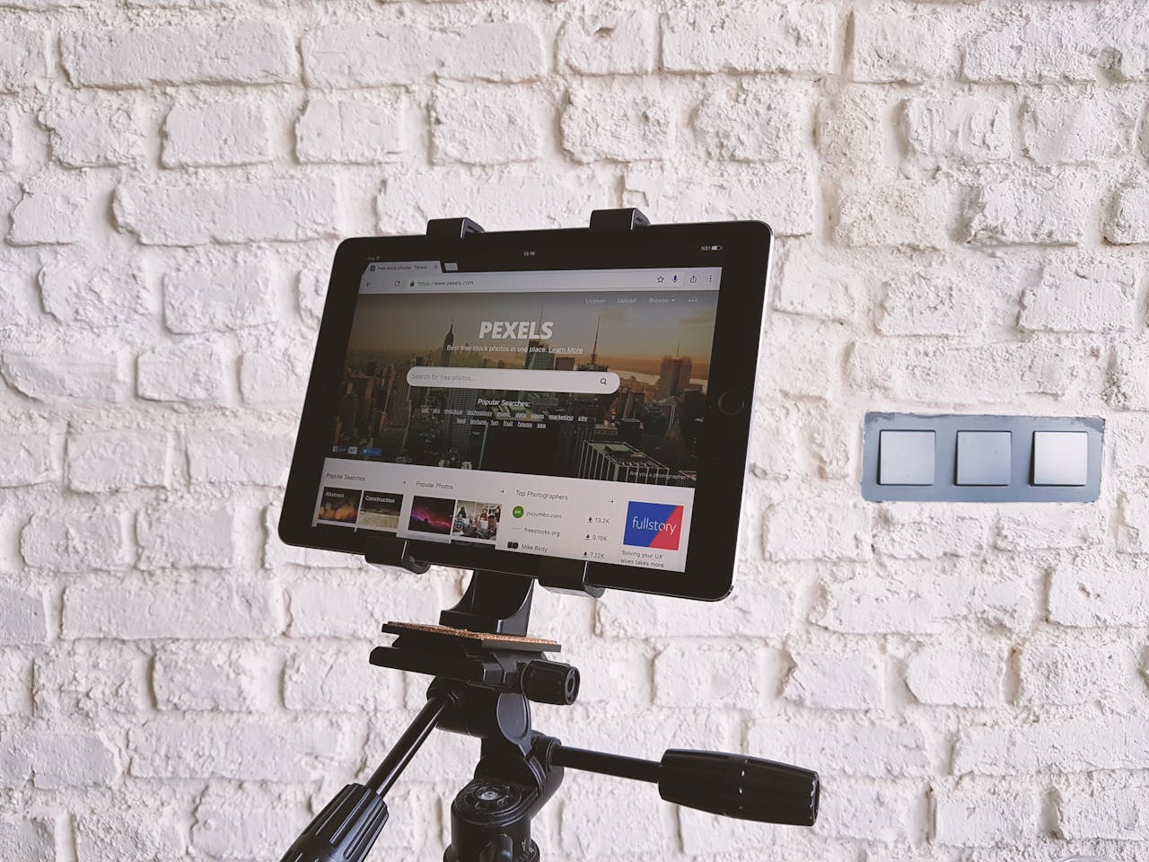 gallery-05 Tablet mounted on a tripod, displaying a website, set against a white brick wall indoors.